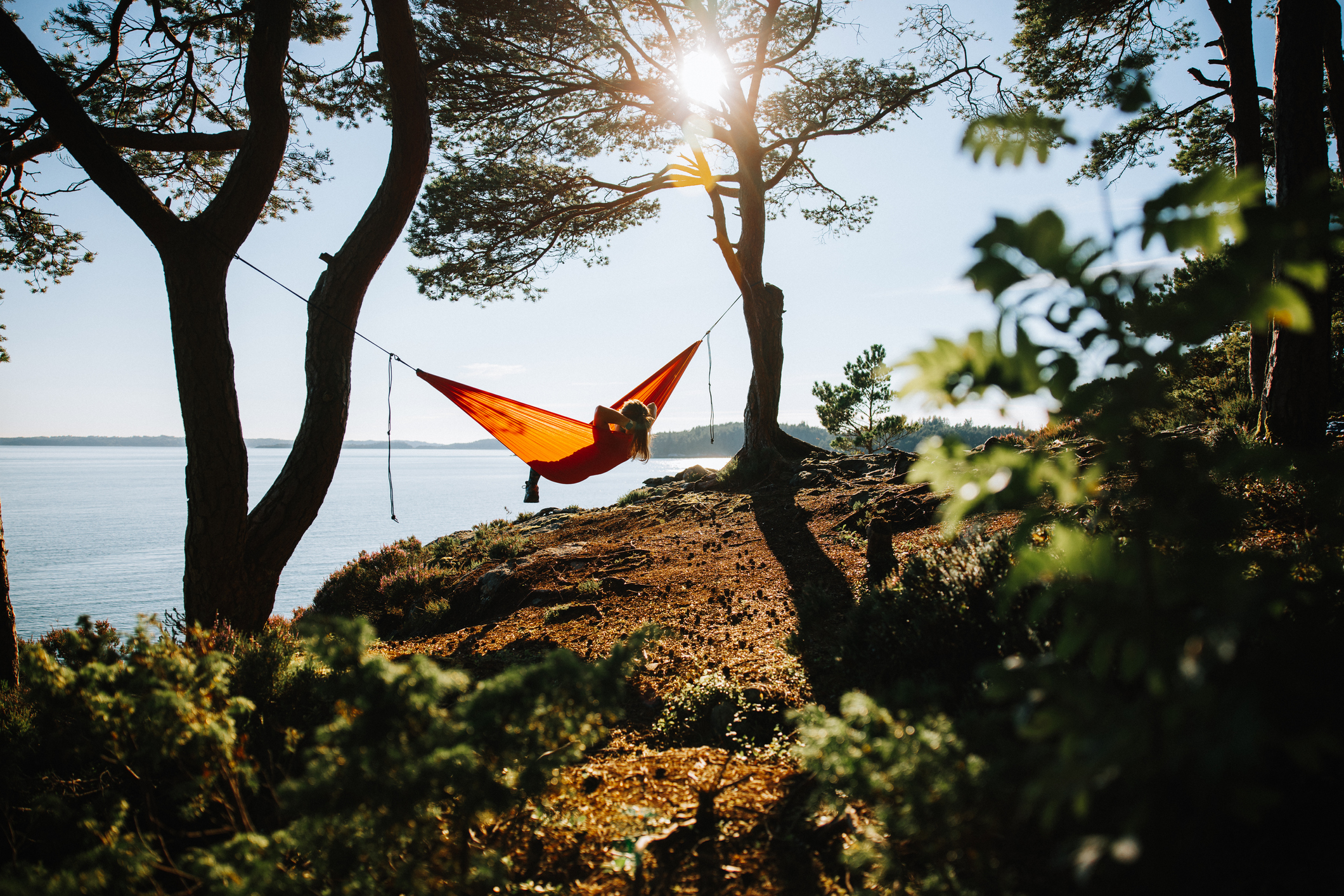 A woman is sitting in a hammock on a rock by the water.